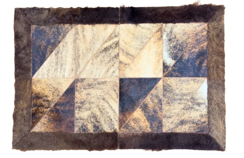 A square patchwork rug featuring a geometric quilted pattern of triangular sections in shades of brown, beige, and dark navy, finished with a thick dark brown fur border.